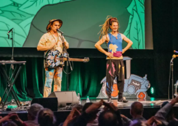 Die Kinderband Rodscha und Tom live auf einer bunten Bühne, umgeben von klatschenden Kindern. MH Eventagentur für Künstlervermittlung und Booking im Emsland und bundesweit.