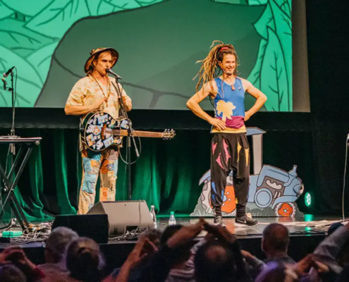 Die Kinderband Rodscha und Tom live auf einer bunten Bühne, umgeben von klatschenden Kindern. MH Eventagentur für Künstlervermittlung und Booking im Emsland und bundesweit.