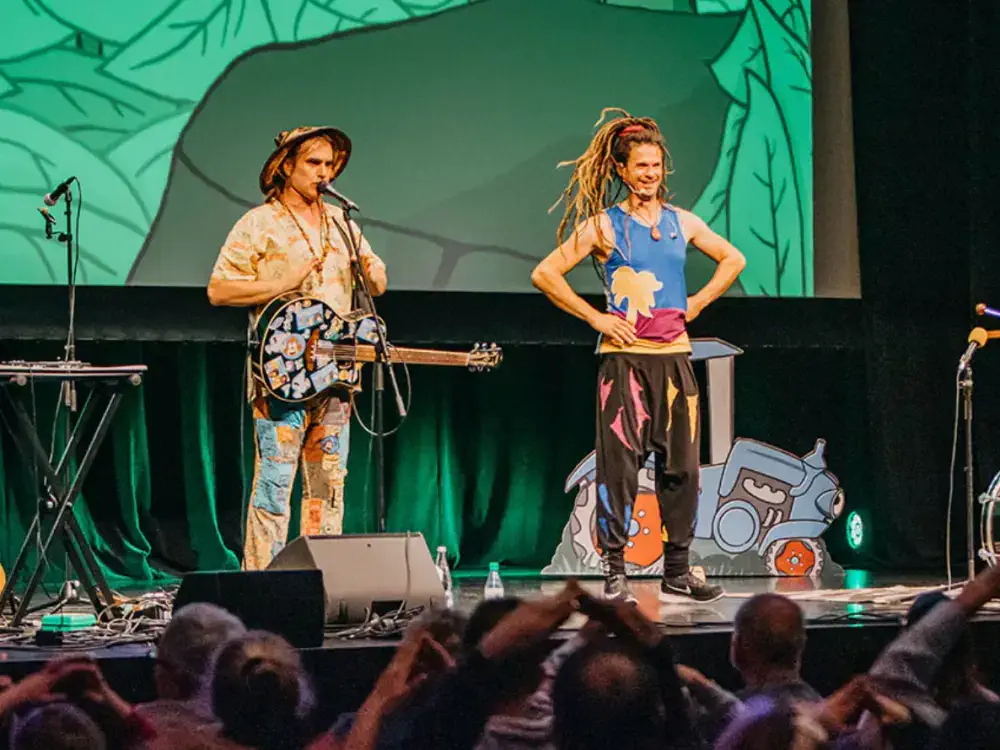 Die Kinderband Rodscha und Tom live auf einer bunten Bühne, umgeben von klatschenden Kindern. MH Eventagentur für Künstlervermittlung und Booking im Emsland und bundesweit.